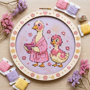 以下が含まれることがあります： ピンク色のバスローブを着た2羽のアヒルが描かれたクロスステッチ刺繍枠。ピンクの花とハートに囲まれています。木製の枠は花とハートで飾られています。ピンク、紫、黄色の刺繍糸と花が枠の周りに配置されています。