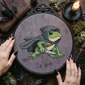 May include: Cross-stitch art of a green frog wearing a hooded cloak and holding a dagger. The frog is centered in a round, dark gray frame. The background is a dark, textured fabric. The frog's design is detailed with various shades of green.