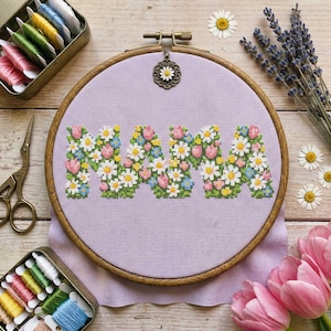 Peut inclure: Un cercle de broderie au point de croix avec le mot "MAMA" brodé en fleurs colorées. La broderie est réalisée sur un tissu lavande, dans un cercle en bois. Fils à broder, ciseaux et fleurs sont disposés autour du cercle.