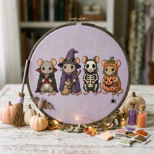 Puede incluir: Un bordado de punto de cruz en un bastidor de madera circular que muestra cuatro ratones con disfraces de Halloween: un vampiro, una bruja con escoba, un esqueleto y una calabaza. El diseño está bordado sobre tela de color morado claro.