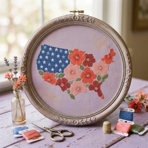 May include: A circular embroidery hoop containing a cross-stitch map of the United States. The western portion features a blue field with white stars, while the remainder is filled with various shades of red, pink, and orange floral patterns.