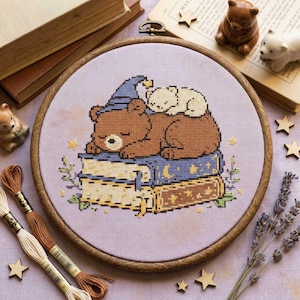 May include: A cross-stitch embroidery in a wooden hoop features a brown bear wearing a blue wizard hat, sleeping atop a stack of books. A smaller white bear rests on top. The design includes stars and floral accents, with embroidery floss and wooden stars nearby.
