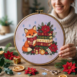 May include: A circular wooden embroidery hoop containing a cross-stitch design on light purple fabric. The pattern features an orange fox wearing a red scarf, sitting beside a stack of books, red poinsettia flowers, and a small gift box.