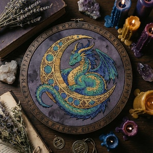 May include: A cross-stitch embroidery of a dragon on a crescent moon. The dragon is green and blue, with gold details. The moon is gold with teal accents, set against a purple backdrop. The embroidery is framed in a wooden hoop with runes.