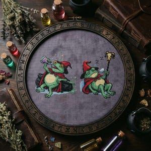 Puede incluir: Bordado de punto de cruz de dos ranas verdes vestidas de magos, con capas y sombreros rojos. Una rana bebe de una botella de poción, la otra duerme con un bastón. El bordado está en un aro de madera con runas.