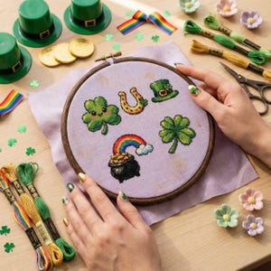 Puede incluir: Un bastidor de bordado con motivos de punto de cruz del Día de San Patricio: un trébol, una herradura, un sombrero de duende, un trébol de cuatro hojas y un arcoíris con un caldero de oro. Hilos de bordar verdes y dorados, sombreros verdes y monedas de oro.