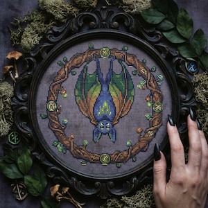 May include: A framed cross-stitch featuring a bat hanging upside down, surrounded by a wreath of branches and leaves. The bat has blue and green wings, and the frame is dark brown. The background is a muted purple.