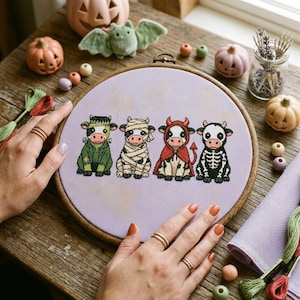 May include: A circular wooden embroidery hoop containing a lavender fabric piece with a cross-stitch design of four cows dressed in Halloween costumes: a Frankenstein monster, a mummy, a red devil, and a skeleton, all arranged in a horizontal row.