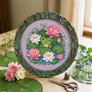 Peut inclure: Une broderie au point de croix encadrée représentant une grenouille verte parmi des nénuphars et des lotus roses et blancs. Le cadre rond a un design décoratif de feuilles. Des nénuphars, des fleurs et des outils de couture sont disposés sur une surface en bois.