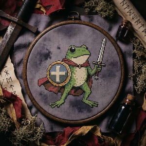 Puede incluir: Bordado de punto de cruz de una rana verde guerrera sosteniendo una espada y un escudo con un diseño de cruz. La rana lleva una capa roja y está sobre un fondo gris dentro de un bastidor de bordado de madera. Accesorios adicionales incluyen una espada, pergaminos y botellas.