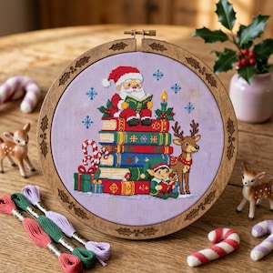 May include: A circular wooden embroidery hoop framing a cross-stitch design on light purple fabric. The image depicts Santa Claus reading a book atop a stack of colorful books, accompanied by a reindeer and an elf. Blue snowflakes surround the scene.