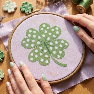 May include: A green and white polka-dotted four-leaf clover cross-stitch design in a wooden embroidery hoop. The clover is on a light purple fabric. Ceramic clovers are on the wooden surface. The image is a close-up of a craft project.