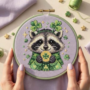 May include: A cross-stitch embroidery hoop featuring a raccoon wearing a green shamrock crown and holding a shamrock. The raccoon is stitched on a lavender fabric, surrounded by green and gold accents. The hoop is held by hands.
