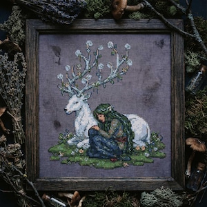 May include: Cross-stitch art in a wooden frame depicts a white deer with elaborate antlers and a person with long green hair. The person is seated, leaning against the deer. The background is a muted purple, with green grass and flowers.