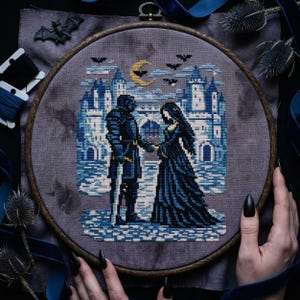 May include: A cross-stitch embroidery featuring a medieval scene with a knight and a woman holding hands in front of a castle. The design uses shades of blue and white, with a crescent moon and bats in the sky. The embroidery is framed in a wooden hoop.