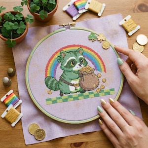 以下が含まれることがあります： 虹の下で金のコインの袋を持った緑のアライグマを描いたクロスステッチ刺繍枠。デザインには、緑と白の市松模様が含まれています。刺繍糸、コイン、小さな鉢植えがフープを囲んでいます。