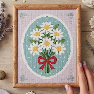 Könnte beinhalten: Kreuzstich-Kunstwerk eines Gänseblümchen-Bouquets mit weißen Blütenblättern und gelben Zentren, gebunden mit einer roten Schleife. Das Kunstwerk ist in einem hellbraunen Holzrahmen mit dekorativen Schnitzereien gerahmt. Der Hintergrund ist ein hellblaues Oval auf lavendelfarbenem Grund.