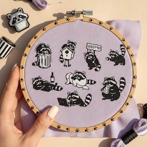Peut inclure: Un cercle de broderie au point de croix avec des illustrations de ratons laveurs noirs et blancs sur un fond lavande. Les ratons laveurs sont représentés dans diverses poses, dont un avec une pancarte « FEED ME! ». Le cercle en bois est tenu par une main.