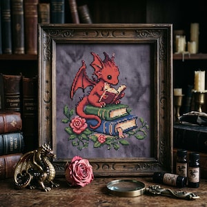 May include: Cross-stitch art of a red dragon reading a book, perched atop a stack of books, surrounded by roses and greenery. The artwork is framed in a dark wooden frame. A bronze dragon figurine and other items are on a wooden surface.