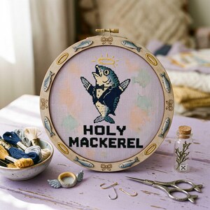 May include: A circular embroidery hoop containing a cross-stitch of a fish wearing a tuxedo with a halo above its head. The text HOLY MACKEREL is stitched in black below the fish. The wooden frame is decorated with small fish and bow motifs.