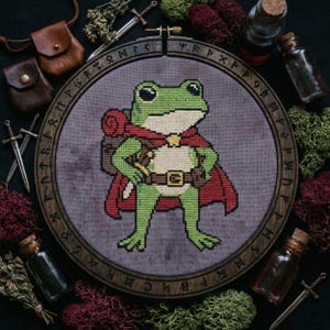 Puede incluir: Bordado de punto de cruz de una rana verde aventurera con capa roja y mochila, en un aro de madera con símbolos rúnicos. La rana tiene un cinturón con hebilla dorada. Pequeñas bolsas de cuero, espadas y botellas rodean el bordado.