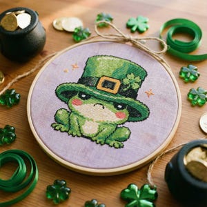 Puede incluir: Un bordado de punto de cruz de una rana verde con un sombrero de duende verde con una hebilla dorada y un trébol de cuatro hojas. La rana está colocada en un aro de bordado de madera. El fondo es de tela morada clara. Monedas de oro y tréboles verdes están esparcidos.