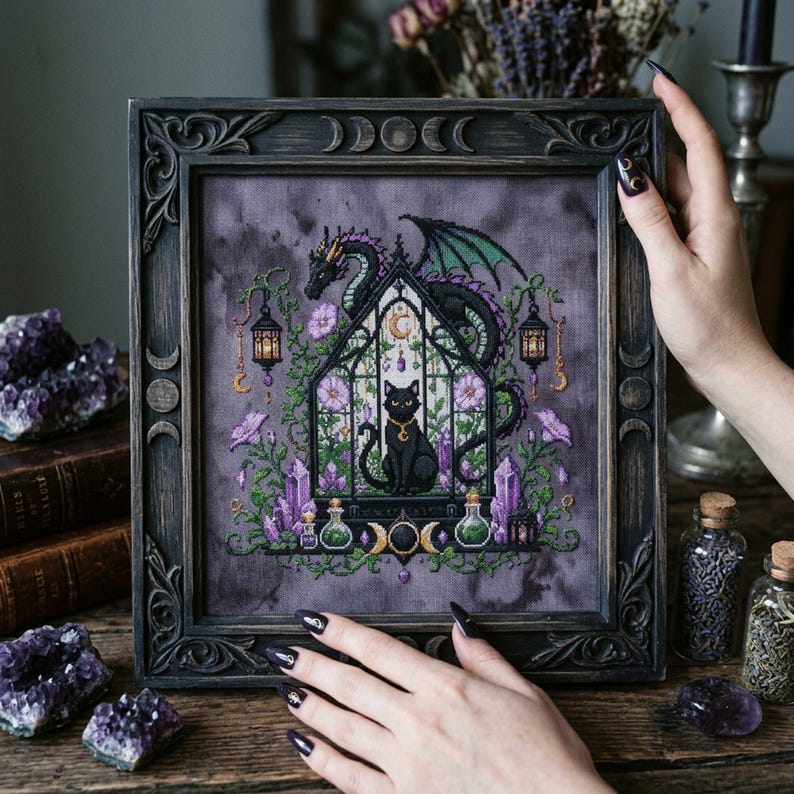magical fantasy cross-stitch pattern featuring a black cat sitting inside a gothic greenhouse while a dark dragon with green wings curls protectively around the roof. The scene is decorated with lavender flowers, leafy vines, purple crystals, potion bottles, hanging lanterns, crescent moon charms, and celestial ornaments. The rich palette of black, green, lavender, gray, purple, and gold creates a mystical witchy garden mood, perfect for cat lovers, dragon fans, fantasy décor, and handmade walL