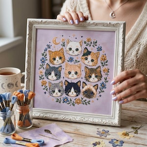 Puede incluir: Obra de arte de punto de cruz enmarcada que presenta nueve caras de gatos de varios colores, dispuestas en círculo, rodeadas de acentos florales azules y amarillos. La obra está sobre un fondo lavanda y enmarcada en un marco blanco ornamentado.