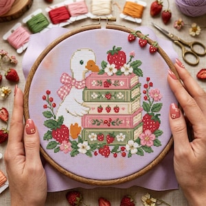 May include: A cross-stitch embroidery project in a round wooden hoop featuring a white duck with an orange beak and a pink checkered bow, standing beside a stack of five books decorated with strawberries and flowers on a light purple fabric.