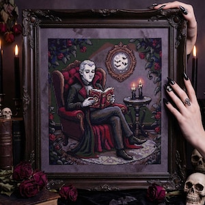 May include: A framed cross-stitch picture shows a vampire reading in a gothic room. The image features a figure in a red chair, holding a book, with a moon and bats in the background. The frame is dark and ornate, with candles and roses.