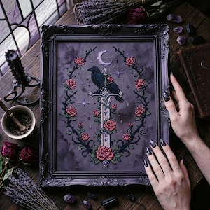 May include: Framed cross-stitch art featuring a raven perched on a sword, surrounded by a wreath of roses and thorns. The artwork includes a crescent moon and stars against a dark gray background. The frame is black.