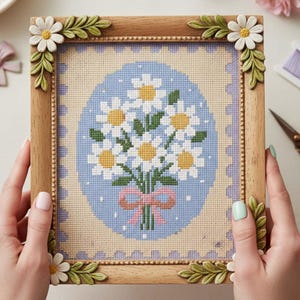 May include: A framed cross-stitch depicting a daisy bouquet with white petals and yellow centres, tied with a pink bow. The artwork is set against a light blue oval background and framed in a wooden frame with daisy and leaf corner accents.