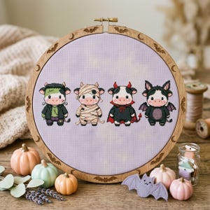 May include: A circular wooden embroidery hoop containing a cross-stitch pattern on light purple fabric. The design features four cartoon cows dressed as a Frankenstein monster, a mummy, a devil, and a bat. The hoop frame has engraved bats.