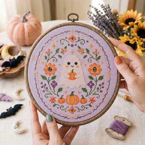 May include: A circular wooden embroidery hoop containing a lavender fabric piece with a cross-stitch design. The pattern features a central white ghost holding a small orange pumpkin, surrounded by orange flowers, green vines, and pumpkins.