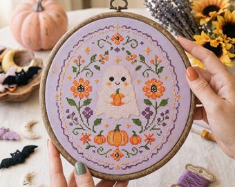 May include: A circular wooden embroidery hoop containing a lavender fabric piece with a cross-stitch design. The pattern features a central white ghost holding a small orange pumpkin, surrounded by orange flowers, green vines, and pumpkins.