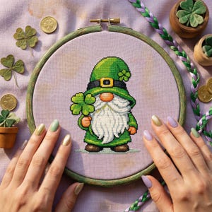 Puede incluir: Bordado de punto de cruz de un gnomo verde sosteniendo un trébol de cuatro hojas, en un aro de madera. El gnomo lleva un sombrero y abrigo verdes con un cinturón negro. Monedas de oro y tréboles están esparcidos sobre una superficie lavanda.