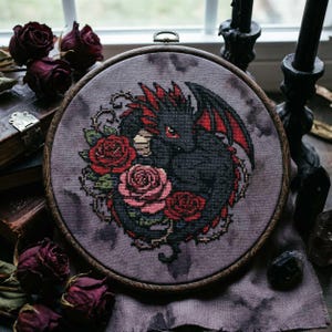 以下が含まれることがあります： 赤とピンクのバラに囲まれた、赤いアクセントの黒いドラゴンを描いた丸いクロスステッチ。ドラゴンは赤い目と翼を持っています。刺繍は、木製のフレームの中で、くすんだ紫色の背景に設定されています。