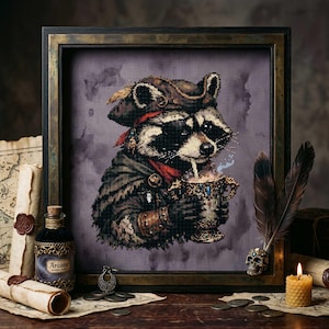 May include: Cross-stitch art of a raccoon pirate, wearing a tricorn hat and red bandana, holding a decorative cup. The framed artwork includes a bottle labeled "Arcane" and a lit candle, with a skull pen holder.