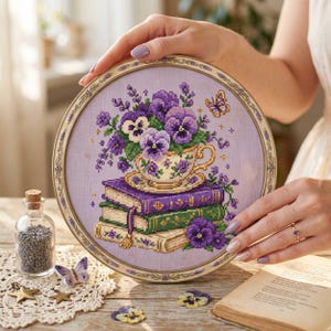 Op de afbeelding: Een borduurring met kruissteek met een bloemenontwerp. Het kunstwerk toont een theekopje vol paarse viooltjes op een stapel boeken. De ring heeft een gouden frame en wordt door een persoon vastgehouden.