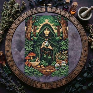 May include: Cross-stitch art of a figure in a green hooded cloak holding a glowing orb, surrounded by forest elements. The artwork is set within a wooden embroidery hoop with etched symbols. Mushrooms, herbs, and potion bottles are also visible.