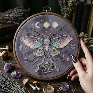 May include: A cross-stitch embroidery featuring a Luna moth design in shades of purple, green, and yellow, set against a gray fabric. The design includes moon phases, stars, and botanical elements, framed in a wooden hoop. The overall theme is mystical and nature-inspired.