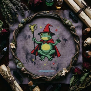 Peut inclure: Point de croix d'une grenouille verte sorcier portant un chapeau et une cape rouges, tenant un bâton. La grenouille est centrée sur un tissu gris, entourée d'une couronne en bois. Thème mystique et sorcier avec des éléments décoratifs.