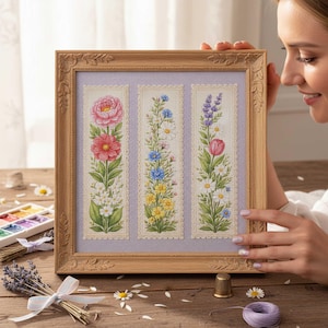 May include: A framed cross-stitch artwork with three vertical panels of floral designs. The panels display a pink peony, mixed blue and white flowers, and a lavender sprig with a pink tulip. The frame is a light brown colour.