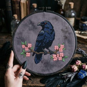 以下が含まれることがあります： 赤い目をした黒いカラスが、ピンクの花が咲く枝に止まっているクロスステッチ刺繍。カラスは灰色の背景に配置されています。刺繍は濃い木製のフープに額装されています。