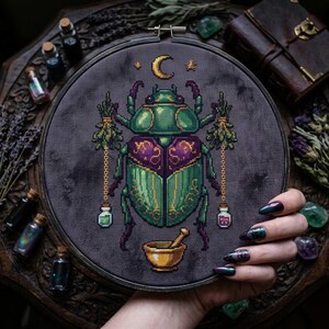 May include: Cross-stitch embroidery in a wooden hoop featuring a green and purple scarab beetle design. The beetle holds sprigs and vials, with a crescent moon and stars above. A golden mortar and pestle are at the bottom. The background is dark gray.