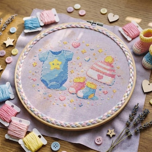 May include: A cross-stitch embroidery hoop featuring a baby-themed design. The design includes a blue onesie with a star, a striped hat, and baby booties. The hoop is surrounded by spools of thread, buttons, and wooden shapes in pastel colors.