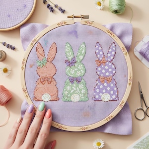 以下が含まれることがあります： パステルカラーのウサギのデザインが3つ施された刺繍枠。桃色、緑色、紫色のウサギにはそれぞれ蝶ネクタイと花の装飾が施されています。刺繍枠はラベンダー色の布の上に置かれ、周りには手芸用品が置かれています。