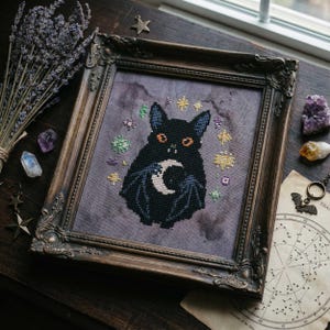 Puede incluir: Arte de punto de cruz de un gato negro con alas de murciélago y una luna creciente, rodeado de estrellas y acentos coloridos. La obra enmarcada tiene un marco de madera oscura con detalles ornamentados. El fondo es morado apagado, con una estética vintage.