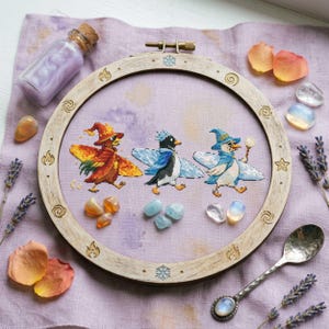 May include: A cross-stitch embroidery hoop featuring three whimsical penguin characters in wizard hats and capes. The penguins are stitched in vibrant colors, set against a lavender fabric. The wooden hoop is decorated with carved designs and surrounded by crystals and dried flowers.