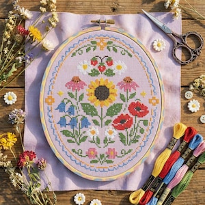 Puede incluir: Un bordado de punto de cruz terminado en un bastidor ovalado de madera. El diseño presenta un vibrante arreglo floral con un girasol, amapolas, margaritas y otras flores en tonos amarillos, rojos, rosas y azules. El fondo es de tela lila claro.